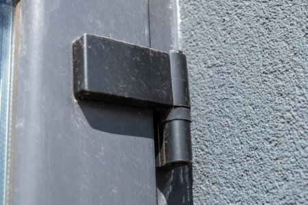 A black door hinge is fixed to a gray wall. The sunlight highlights the details of the hinge. Dust is visible on its surface.