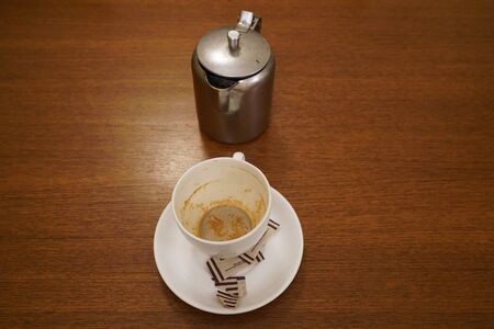 Old dirty used aluminum tea pot kettle with dirty used white tea mug. Antique Aluminum Kettle with tea cup and empty used sugar sachets. Dirty coffee and tea cup half empty on saucer on table.の写真素材