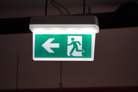 Glowing Emergency exit sign with left arrow at a building. Safety first concept. Copy space wallpaper. Exit signs with light in a parking of building.の写真素材