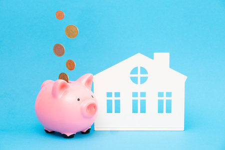 Coins are falling into a pink piggy bank near the house on a blue background. The concept of...