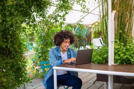 Smiling young ethnic guy 20 years old, dressed in casual clothes, works on a laptop outdoors, in a green area in nature. Green open space for coworking, freelancer, relaxing, coffee, food. Free place to work, business, study, freelance in natureの写真素材