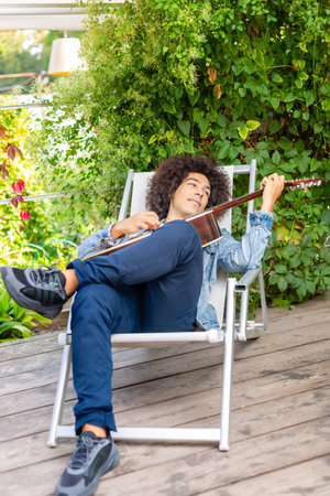 Beautiful young Afro-Italian mixed race man in casual wear playing guitar in the garden. A multiracial boy plays a musical instrument rests and relaxes outdoors. Spending time at homeの写真素材