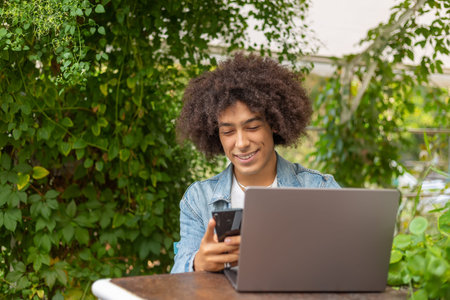 Smiling young ethnic guy 20 years old, dressed in casual clothes, works on a laptop outdoors, in a...