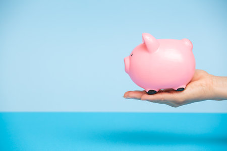 Person puts money in a pink piggy bank on a blue background. Women's hands put coins in a money box.Piggy bank with stacks of coins.The concept of investment in the future, savings, financial planning.の写真素材