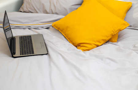 Workplace in bed. Laptop with glasses on the bed on gray bedding and yellow pillows. Modern and comfortable bedroom, home office background. The concept of working from home.の写真素材