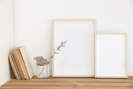 Empty wooden frame on white wall background with glass vase and green eucalyptus branches on wooden table with books Neutral modern organic still life interior. Poster mockupsの写真素材