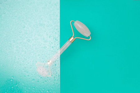 Pink jade roller for facial aesthetic massage on turquoise water background with water drops. Skin...