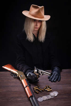 wild west blonde girl with gun at the table with ammunition and silver coins on black backgroundの写真素材