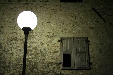 Old streetlight that illuminates a wall with a window at nightの写真素材