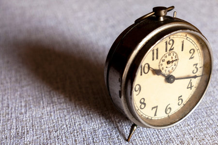 Old alarm clock, metal clock, with white dial with numbers and black hands over a table that casts a...