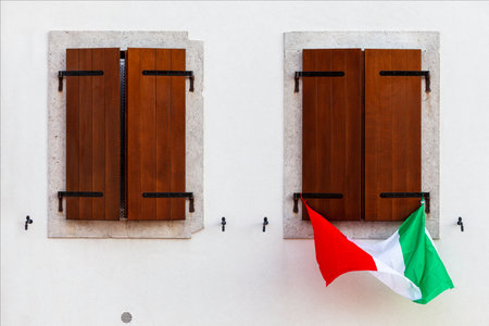 Italian flag hanging from one of the two closed windows with brown wooden balconies. Building facade in white color style. Historic center of the fortress city of Palmanova.の写真素材