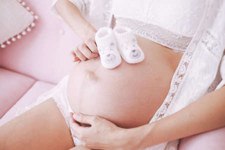 Closeup of pregnant woman sitting and touching her belly, with baby shoes on her belly.の写真素材