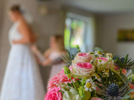 Beautiful wedding colorful bouquet for bride in foreground and bridesmaid is helping the bride to dress at blur background. Beauty of colored flowers. Close-up bunch of flowers. Bridal accessories.の写真素材