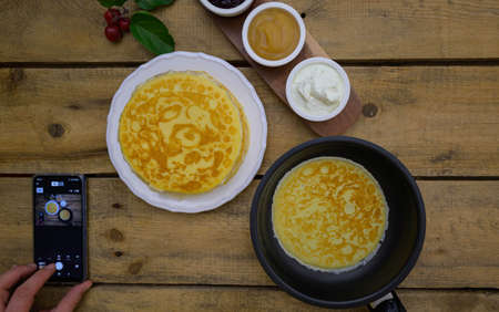 Remote control with help of smartphone by photo shooting of pancakes. Russian pancakes crepes in stack and on the pan with apple sauce and sour cream on rustic wooden background.の写真素材