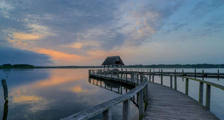 Swinging curved pier construction and shelter with thatched roof at lake Hemmelsdorf, Schleswig-Holstein, Northern Germany. Winding wooden footbridge and bird platform on the lakeの写真素材