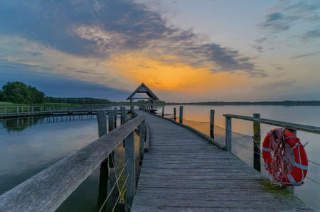 Swinging curved pier construction and shelter with thatched roof at lake Hemmelsdorf, Schleswig-Holstein, Germany. Winding wooden footbridge, bird platform and lifebelt on the lakeの写真素材