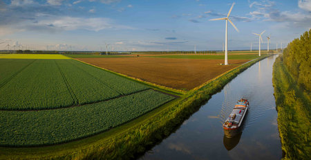 Agricultural land along canal with boat moving on canal and row of rotating wind turbines along bank of canal. Onshore with wind turbines on agricultural land.の写真素材