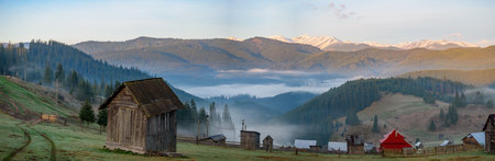 View of mountain village surrounded by peaks of Carpatian mountains with wooden cabins and faint trail of an automobile tires on dews grass. Mist gently blankets mountain valley viの写真素材
