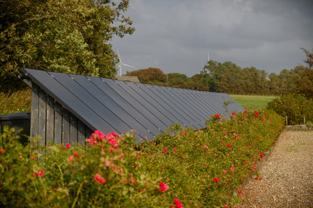 Panorama view of solar panels on the roof of shed, set amidst blooming shrubs with wind turbines partially visible behind trees. Solar panels on the roof of garbage can shelter.の写真素材