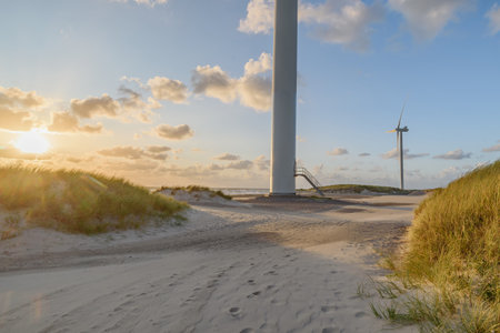 Coastal sunset with wind turbines on sand dunes with dune grass. Metal staircase leads up to a door on the wind turbine, surrounded by dune grass and footprints heading towards theの写真素材