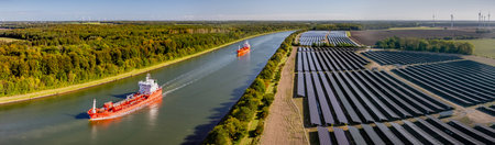 Tanker transporting fossil fuels along waterway, sailing past canal bordered by solar farm. Traditional and modern energy. Concept of transition from fossil fuels to renewable energyの写真素材
