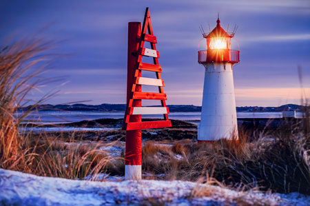 Coastal lighthouse glowing warmly against winter twilight, with striking red and white triangular navigational marker beside it, set amidst snow-dusted dunes, frosty grasses and puの写真素材