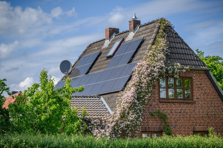 Solar-equipped residential brick house with a roof fitted with photovoltaic panels and a facade partially covered by a flowering Clematis vine.の写真素材