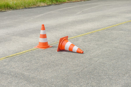 Two orange traffic cones on a paved road, one upright and one tipped over with a yellow line running...