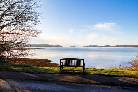 An empty wooden seat or bench over looking the sea at Kirkcudbright Bay, during low tide on a sunny winter morning, Dumfries and Galloway, Scotlandの写真素材