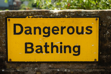 A yellow dangerous bathing warning sign fixed to a stone wall beside waterの写真素材