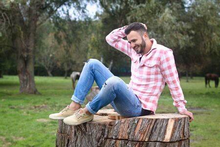 Young handsome guy Cowboy. farmer is sitting on a large stump on his ranch. Rural landscapes, countryside. Trees, field, farm. Stock photos for designの写真素材