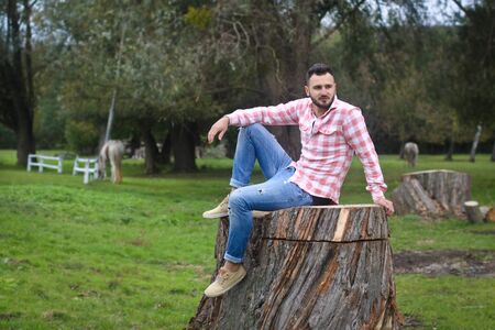 Young handsome guy Cowboy. farmer is sitting on a large stump on his ranch. Rural landscapes, countryside. Trees, field, farm. Stock photos for designの写真素材
