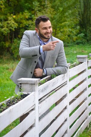 Portrait of a sexy man dressed in a suit. Fashionable guy in the autumn nature near a white wooden fence. Businessman relaxes. Skok photo for designの写真素材