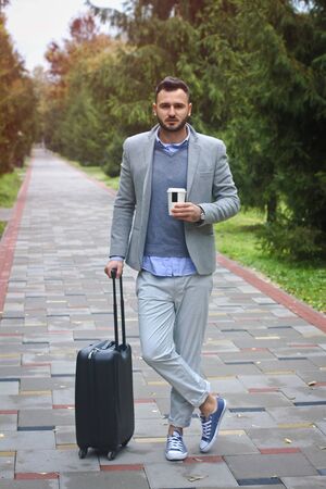 Fashionably dressed guy is traveling with luggage in the background of a road and trees. The man hurries on his flight and pours coffee, tea. Stock photo for designの写真素材