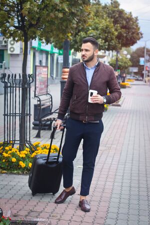 Handsome man with a suitcase for tourism. Exit the house for travel. Fashionably dressed guy and coffee. Stock Photo.の写真素材