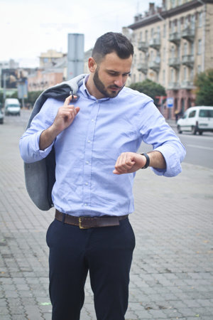 Portrait of a guy in a shirt sitting in a park in a European city. A man looking at the clock against the background of trees and buildings. Stock photosの写真素材
