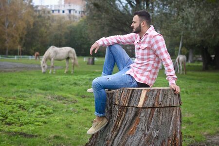 Young handsome guy Cowboy. farmer is sitting on a large stump on his ranch. Rural landscapes, countryside. Trees, field, farm. Stock photos for designの写真素材