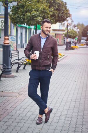 Man in a jacket and suit on a European street with coffee in his hands. Business guy fashion clothes. Autumn landscape, portrait. stock photoの写真素材