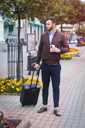 Handsome man with a suitcase for tourism. Exit the house for travel. Fashionably dressed guy and coffee. Stock Photo.の写真素材