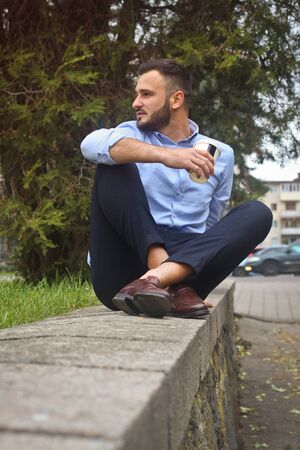 Portrait of a guy in a shirt sitting in a park in a European city. A man in the background of trees and buildings. Stock photosの写真素材