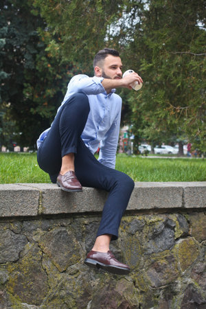 Portrait of a guy in a shirt sitting in a park in a European city. A man in the background of trees...