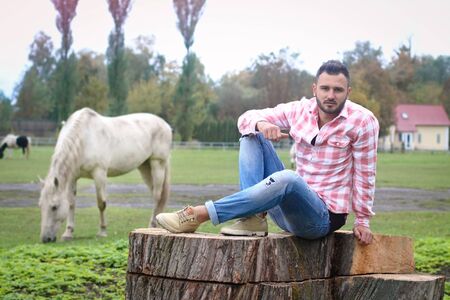 Young handsome guy Cowboy. farmer is sitting on a large stump on his ranch. Rural landscapes, countryside. Trees, field, farm. Stock photos for designの写真素材