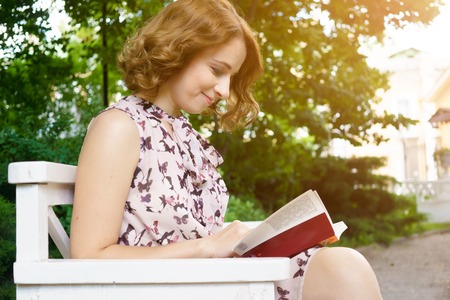 Beautiful young woman reading book on park bench on summer sunny day. Image with lens flare effectの写真素材