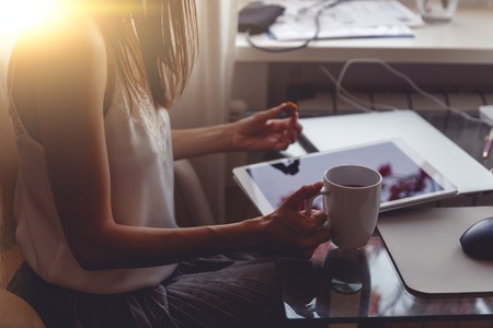 Brunette girl sitting at the table, holding cup with hot drink and tablet on table. Image with lens flare effect and split tonedの写真素材