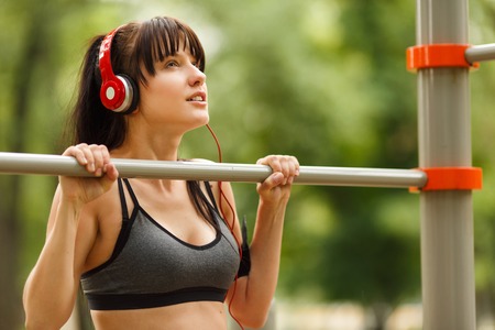 Young brunette woman listening to music and doing exercises on the horizontal bar in park on summer dayの写真素材
