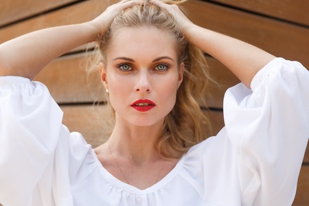 Gorgeous blonde woman with natural makeup and red lips in white blouse. Portrait on background of wooden wallの写真素材