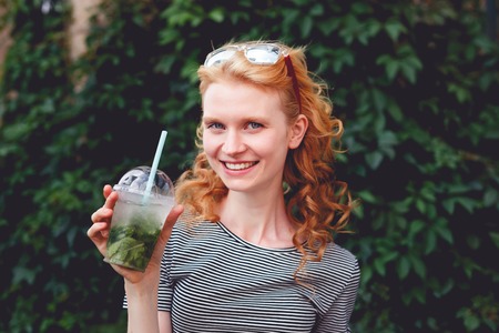 Smiling ginger woman in sunglasses with cocktail in hand at green bush, photo tonedの写真素材