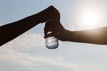 Two people holding bottle of water on background sky and sun. Two outstretched hands closeupの写真素材