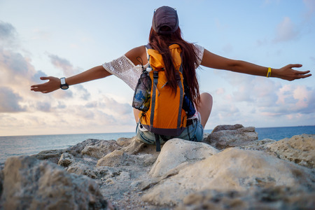 Young sports woman tourist sitting on rock, resting and looking at sea. Freedom camping concept. Model in summer clothes, peaked cap and travel backpackの写真素材