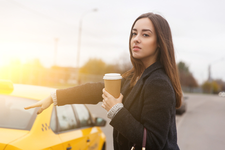 Thoughtful brunette with coffee stoping yellow taxi on streetの写真素材